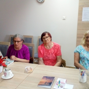 Wnętrze biblioteki, cztery seniorki siedzące przy stole i dyskutujące podczas spotkania klubu "Seniorzy z książką na ty".