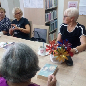 Wnętrze biblioteki, trzy seniorki siedzące przy stole i dyskutujące podczas spotkania klubu "Seniorzy z książką na ty".