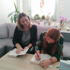 Magdalena Kordel oraz Wioletta Wachewicz - Sekretarz Powiatu Łęczyńskiego siedzące na kanapie, autorka wpisuje dedykację do książki.