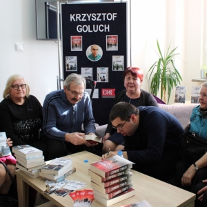 Krzysztof Goluch oraz sześciu członków z DKK w Ludwinie siedzących na kanapach, autor wpisuje dedykację do książki.